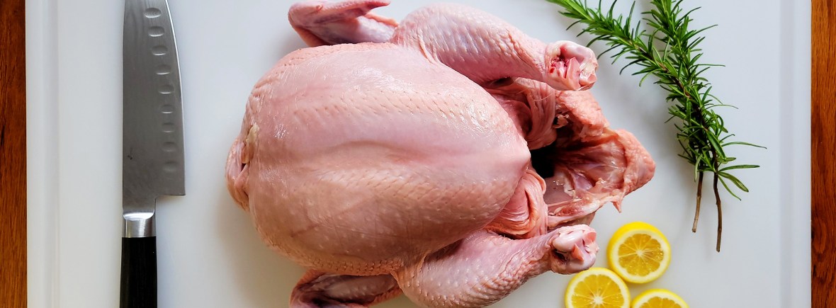 Whole Chicken carcass on cutting baord with rosemary, a chefs knife, and lemon slices