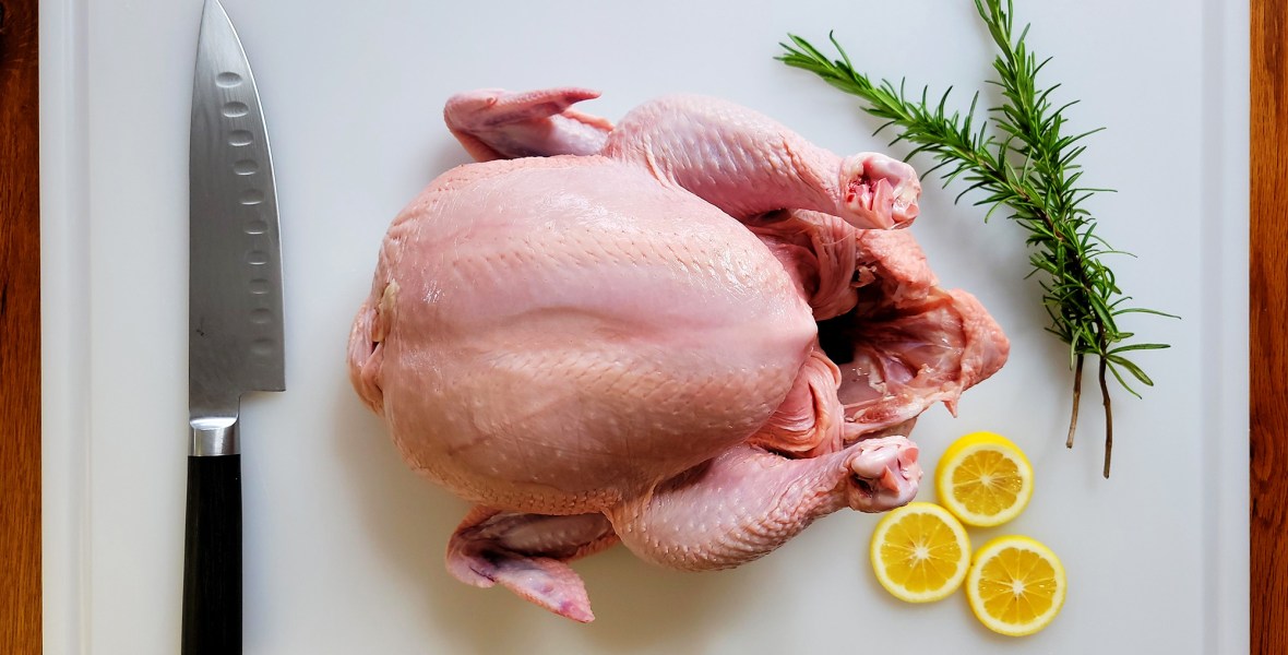 Whole Chicken carcass on cutting baord with rosemary, a chefs knife, and lemon slices